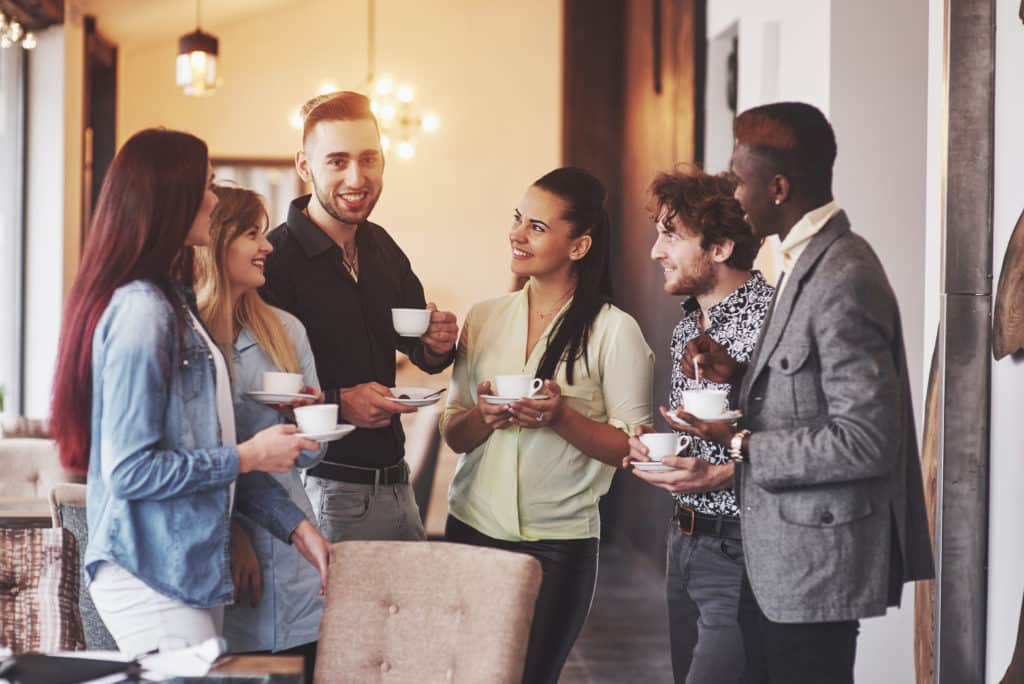 Six personnes ont un café à la main, discutent et sourient aux autres.